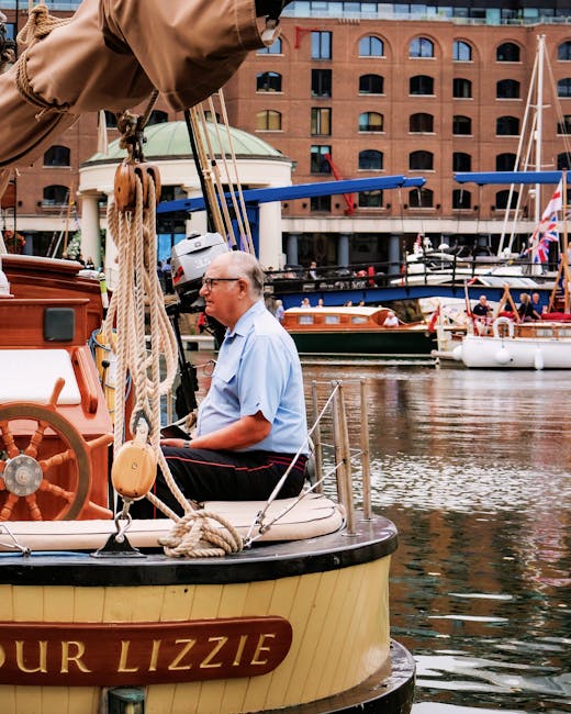 The image features a large, historic sailing ship moored at a marina near a modern urban area. The ship has a black hull and white sails, some of which display a red cross emblem. The sails are furled
