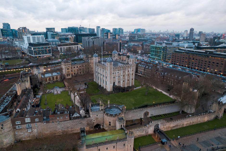 A view of the London skyline featuring the Shard skyscraper towering over modern office buildings with glass facades. In the foreground, there are green trees and part of a blue and white railing stru
